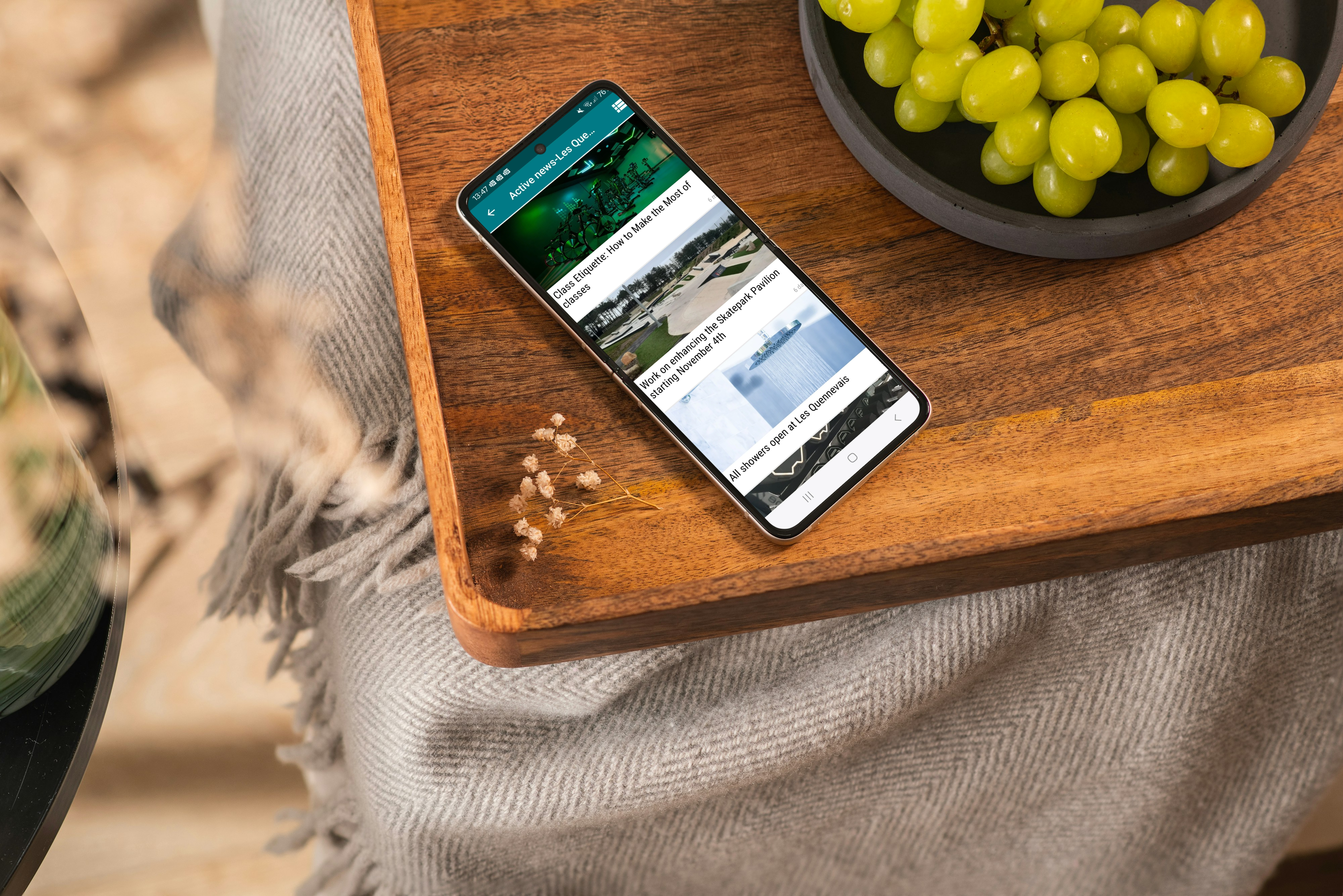 photo of the news section of the active app open on a mobile phone that is placed on a wooden table with a bowl of grapes next to it